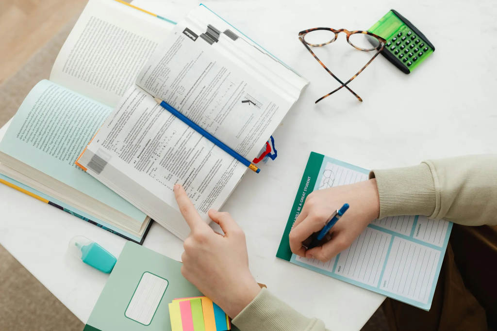 Textbooks, glasses, a highlighter, and a calculator are laid out on a desk. Two hands are visible, left hand pointing at a text book, right hand is making annotations on a worksheet.